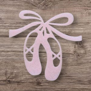 May include: A light pink felt cutout shaped like a pair of ballet slippers tied together with a decorative bow. The design features cut-out details representing the shoe openings and crisscross ribbon lacing on the front of each slipper.