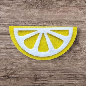 May include: A semi-circular felt cutout shaped like a lemon slice, featuring a bright yellow outer rind and a white inner pith section with five distinct triangular segments radiating from the center point of the flat edge.