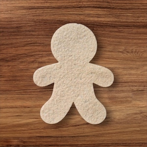 Op de afbeelding: Een beige vilten peperkoekmannetje, met uitgestrekte armen en benen, ligt op een bruin houten oppervlak. Het vilt heeft een textuur. De afbeelding is een bovenaanzicht.