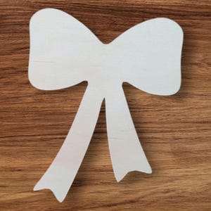 May include: A white wooden bow-shaped cutout, featuring a classic bow design with two long, downward-pointing tails. The bow is set against a warm, brown wood grain background, creating a contrast in texture and color.