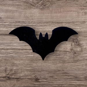 Può includere: Una sagoma piatta e nera di un pipistrello con le ali spiegate. Il pipistrello presenta due orecchie appuntite sulla sommità della testa e una sezione centrale del corpo che si restringe verso il basso, creando una forma simmetrica.