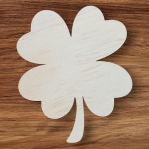 May include: A light-coloured wooden shamrock cutout with four heart-shaped leaves and a stem. The shamrock is set against a brown wood grain background, creating a natural and rustic aesthetic. Suitable for St. Patrick's Day decorations or craft projects.