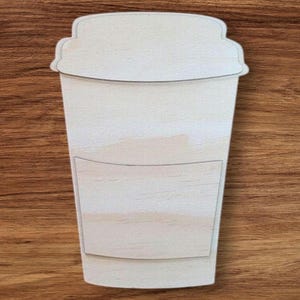 May include: A wooden cutout in the shape of a coffee cup. The cup has a lid and a sleeve detail. The wood has a natural, light color and is set against a brown wooden surface. This item is suitable for crafts and DIY projects.