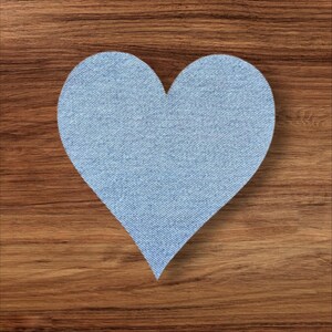 May include: A light blue denim heart shape on a wooden surface. The heart is cut from denim fabric, showing the typical diagonal weave. The background is a warm-toned wood grain.