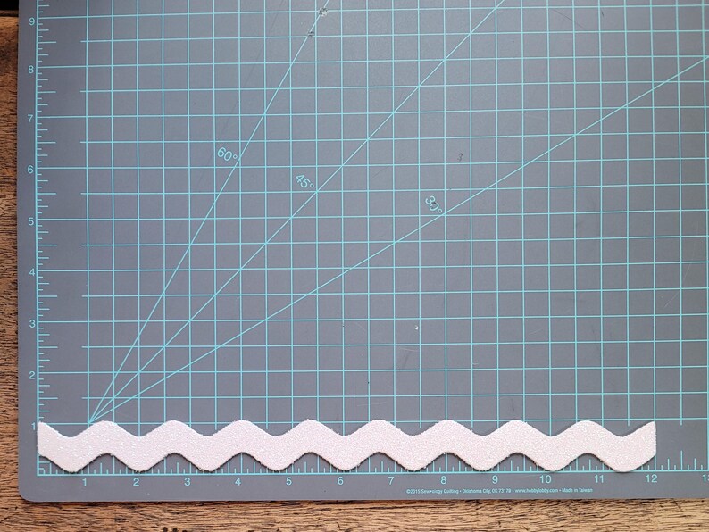 May include: White zig zag trim on a gray cutting mat with a grid pattern. The trim is about 12 inches long.