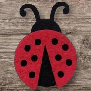 May include: A red and black felt ladybug with black spots and antennae. The ladybug is a decorative item, possibly a craft or embellishment, with a simple, stylized design. The background is a wooden surface.