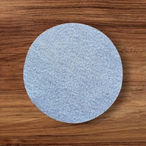 May include: A round coaster with a denim fabric design. The coaster is light blue and has a textured appearance. The coaster is placed on a wooden surface.