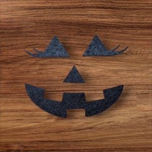 May include: A set of dark charcoal gray felt cutouts forming a jack-o'-lantern face. The pieces include two triangular eyes with curved eyelashes, a small triangular nose, and a wide, jagged mouth with two square teeth in the center.