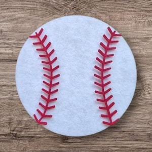 Op de afbeelding: Een cirkelvormige witte viltuitsnede in de vorm van een honkbal, voorzien van twee gebogen lijnen van rood vilt. De rode stiksels zijn gerangschikt in een klassiek honkbalpatroon met kleine, schuine kruissteken langs elke gebogen lijn.
