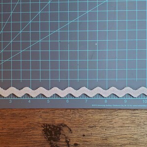 May include: A white, wavy trim with a glittery finish. The trim is approximately 12 inches long and is shown on a green cutting mat with inch markings.