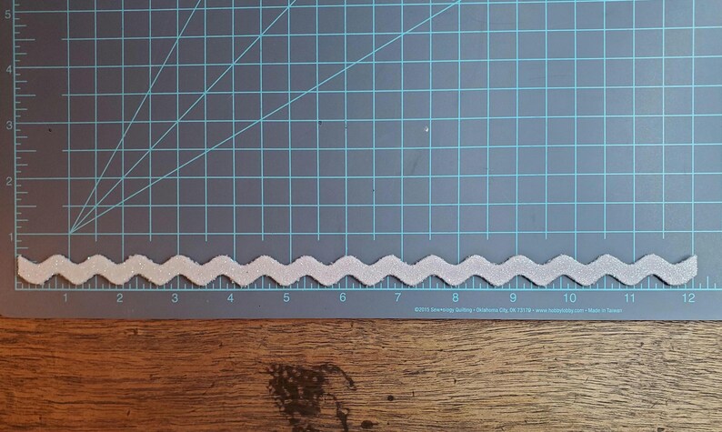 May include: A white, wavy trim with a glittery finish. The trim is approximately 12 inches long and is shown on a green cutting mat with inch markings.