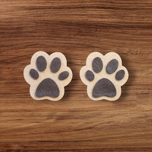 May include: Two beige paw print coasters with dark gray paw pads are displayed on a wooden surface. The coasters are made of a soft material and are designed to protect surfaces from scratches and spills. These coasters are perfect for pet lovers.