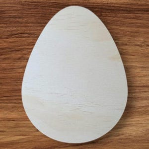 May include: A light-colored, wooden egg-shaped cutout, likely for crafting or decoration. The egg is smooth and unpainted, with visible wood grain. It sits on a brown wooden surface.
