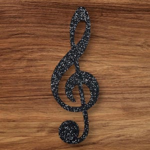 May include: A black glitter treble clef music note cutout. The note is on a wood background.