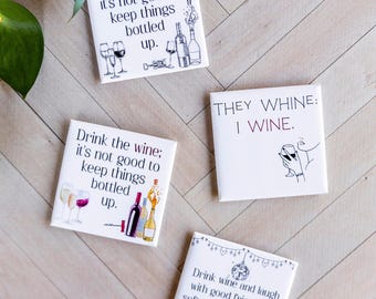 May include: Four white square coasters with wine-themed quotes and illustrations. The coasters feature phrases like "Drink the wine; it's not good to keep things bottled up" and "They whine: I wine."
