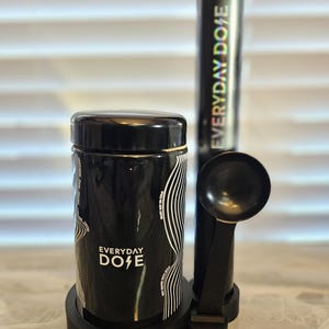 May include: A black ceramic container with the text "EVERYDAY DOSE" in white, accompanied by a black scoop and a tall, cylindrical container. The containers have a white wave design. The items are on a black base.