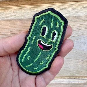 May include: A green embroidered patch in the shape of a smiling pickle with a black border. The pickle has a happy face with white eyes, a pink mouth, and a green body. The patch is held in a hand.
