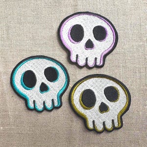 May include: Three embroidered skull patches with black eye and nose sockets. Each skull has a white base with a colored outline: teal, purple, and gold. The patches are on a neutral linen background.