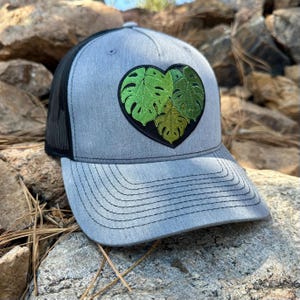 May include: A gray baseball cap with a black mesh back and a curved brim. The cap features a heart-shaped patch with green monstera leaves. The patch is outlined in black.