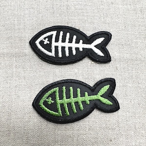 May include: Two embroidered patches in the shape of a fish skeleton. One patch has a black background with white stitching, and the other has a black background with green stitching. The patches are likely for clothing or accessories.