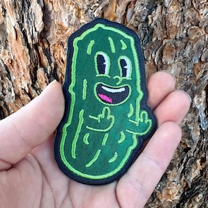 May include: A green embroidered patch featuring a cartoon pickle character with a smiling face and raised middle fingers. The patch has a black border and is held against a textured brown background.