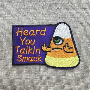 May include: Embroidered patch featuring a candy corn character giving the middle finger. The patch has a purple background with the words "Heard You Talkin Smack" in orange. The candy corn has a white top, orange middle, and yellow bottom.