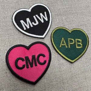 May include: Three heart-shaped embroidered patches. One is black with white text "MJW", another is pink with black text "CMC", and the third is green with gold text "APB". Each patch has a contrasting border.