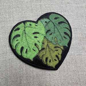 May include: Heart-shaped embroidered patch featuring three green Monstera leaves on a black background. The leaves have varying shades of green, with detailed stitching. The patch is designed for application on fabric items.