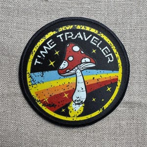May include: A black and yellow circular patch with the text "TIME TRAVELER" in a white circle. A red and white mushroom with white spots is in the center of the patch, with a rainbow stripe behind it.