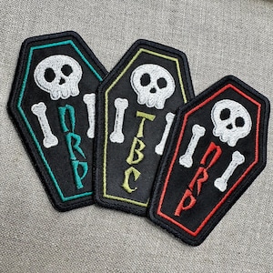 May include: Three black coffin-shaped patches with white skull and crossbones designs. Each patch has a different colored border and text: teal with "NRP", yellow with "TBC", and red with "DRP".
