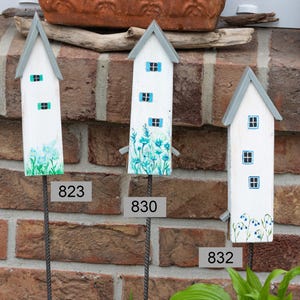 Könnte beinhalten: Drei weiße, hausförmige Gartenstecker aus Holz mit grauen Dächern und blauen Fensterdetails. Jeder Stecker hat bemalte Blumenakzente und ist auf einem Metallstab montiert. Die Nummern 823, 830 und 832 sind aufgedruckt.
