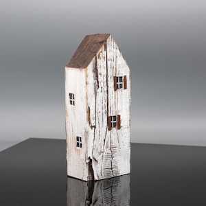 Könnte beinhalten: Eine hohe, schmale Holzhaus-Skulptur mit verwitterter weißer Lackierung und einem Dach aus naturbelassenem braunem Holz. Das Objekt zeigt mehrere kleine, handgemalte schwarze Fenster und zwei braune Fensterläden auf der Vorderseite.