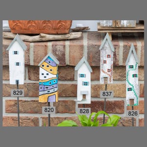 Könnte beinhalten: Eine Sammlung handbemalter Holzhausdekorationen. Jedes Haus hat ein einzigartiges Design mit verschiedenen Farben, Fensterdetails und Dachstilen. Die Häuser sind auf Metallstäben montiert, ideal für Garten- oder Hofdekoration. Maße in cm.