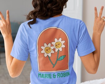 Puede incluir: Camiseta azul claro con un diseño retro de dos flores de margarita con ojos en forma de corazón dentro de un arco naranja. El texto "MARIE & ROBIN" está impreso en verde debajo. La camiseta es de material suave.