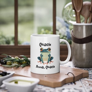 Puede incluir: Taza de cerámica blanca con un diseño de rana de dibujos animados, con ojos naranjas y detalles azules y verdes. La taza tiene el texto "Quack Anna, Quack" en negro. La taza está sobre una tabla de cortar de madera.
