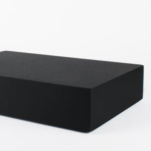 Könnte beinhalten: Ein schwarzer, rechteckiger Block mit strukturierter Oberfläche vor weißem Hintergrund. Die Form des Blocks ist schlicht und modern. Die Abmessungen betragen ca. 30 cm Länge und 15 cm Breite.