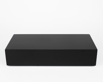 May include: A solid black rectangular box with a matte finish. The box is a simple, modern design, with clean lines and a smooth surface. The object is set against a plain white background.