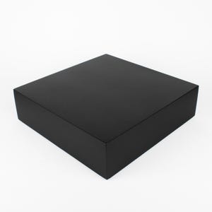 Könnte beinhalten: Eine quadratische, schwarze Box mit mattem Finish. Die Box ist einfarbig und hat ein schlichtes, modernes Design. Die Box wird wahrscheinlich für Verpackungs- oder Ausstellungszwecke verwendet.