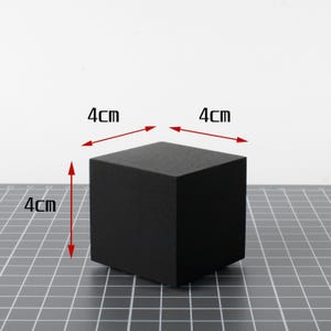 May include: A solid black cube, each side measuring 4cm. The cube is displayed on a grid surface, with arrows indicating the dimensions. The object is a simple geometric shape.