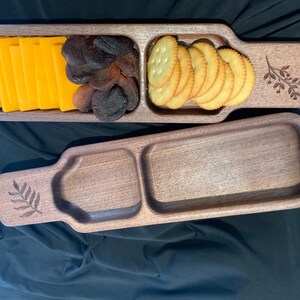 May include: A wooden serving tray with three compartments. The tray is made of dark wood and has a natural finish. The compartments are filled with snacks, including cheese slices, dried fruit, and crackers.