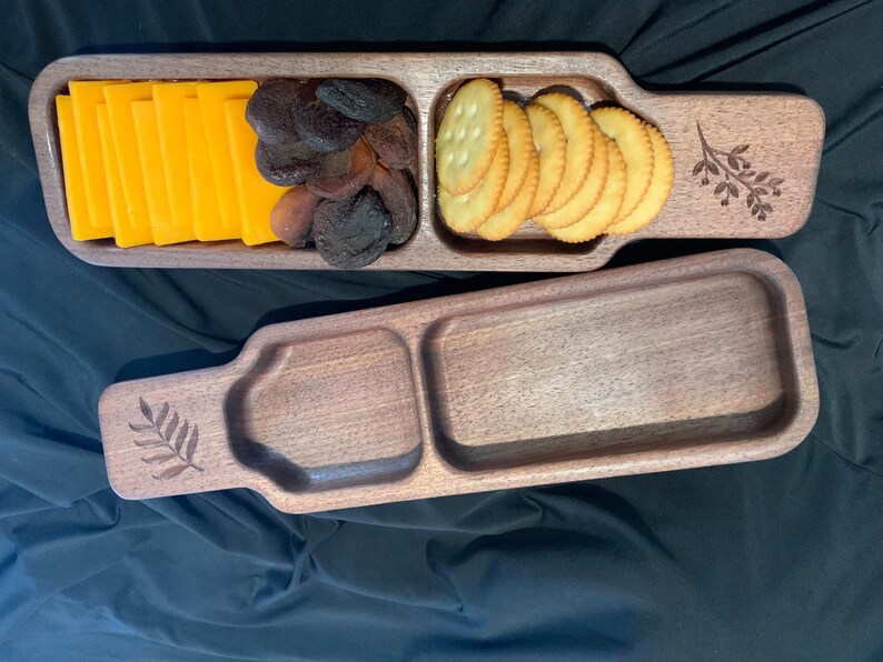 May include: A wooden serving tray with three compartments. The tray is made of dark wood and has a natural finish. The compartments are filled with snacks, including cheese slices, dried fruit, and crackers.
