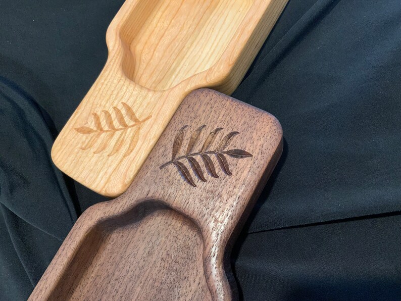 May include: Two wooden serving trays with a carved leaf design. The tray on top is light wood and the tray on the bottom is dark wood.