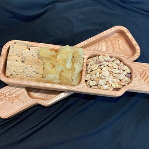 May include: A wooden serving tray with three divided sections. The tray is made of light brown wood and has a carved leaf design on the handle. The tray is filled with snacks, including crackers, potato chips, and peanuts.