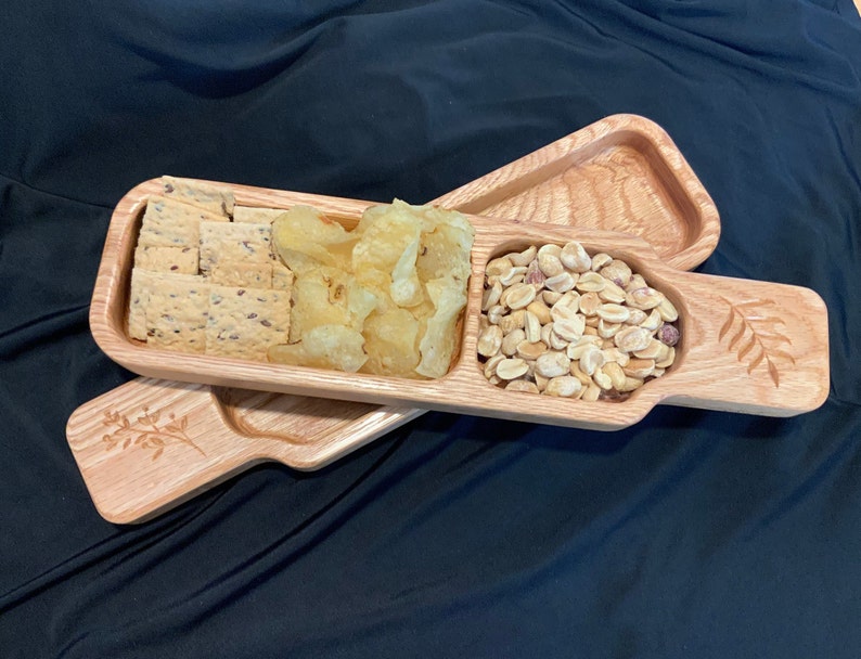 May include: A wooden serving tray with three divided sections. The tray is made of light brown wood and has a carved leaf design on the handle. The tray is filled with snacks, including crackers, potato chips, and peanuts.