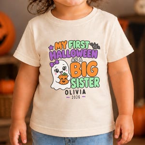 May include: A natural-coloured t-shirt featuring a graphic with the text "MY FIRST HALLOWEEN AS A BIG SISTER" above the name "OLIVIA" and the year "2026". The design includes a white ghost holding a pumpkin, a purple bat, and stars.