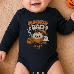May include: A black long-sleeve baby bodysuit featuring a graphic of a bao bun in a bamboo steamer, a ghost, and a jack-o'-lantern. The text reads PUMPKIN BAO, NOAH, and 2026. The design includes colorful candy and small ghost illustrations.