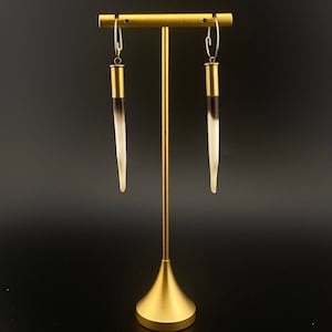 May include: A pair of long, black and white earrings with gold accents hanging from a gold stand.