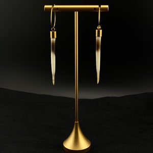 May include: A pair of gold hoop earrings with a white and brown horn pendant hanging from each hoop. The earrings are displayed on a gold stand against a black background.