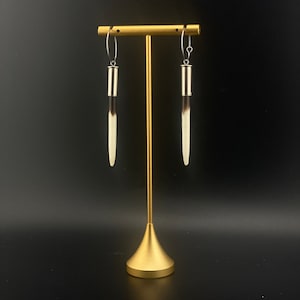 May include: A pair of long, white and black earrings hanging from a gold stand. The earrings are made of a natural material, possibly bone or horn.