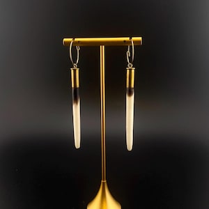 May include: A pair of long, white and black earrings with gold accents hanging from a gold stand against a black background.